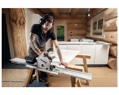 Un artisan avec des lunettes de protection et un casque antibruit scie une plaque avec une scie plongeante Metabo dans une pièce aux murs en bois.