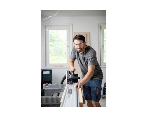 Un homme scie une planche de bois avec une scie circulaire portative sur un rail de guidage, tout en portant des lunettes de protection.