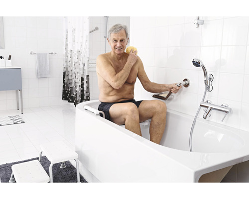 Ein älterer Mann benutzt eine Badewanne mit Einstiegshilfe und Haltegriff in einem hellen Badezimmer.