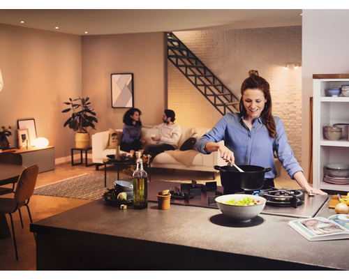 Cuisine d''habitation ouverte et lumineuse avec une femme cuisinant à la cuisinière et des personnes assises sur le canapé en arrière-plan.