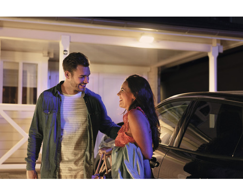 Un couple se sourit devant une maison et une voiture.