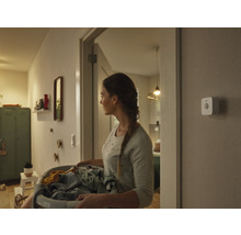 Une femme porte un panier à linge dans un couloir, un détecteur de mouvement intelligent est fixé au mur.