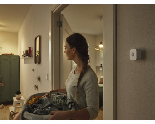 Une femme porte un panier à linge dans un couloir, un détecteur de mouvement intelligent est fixé au mur.
