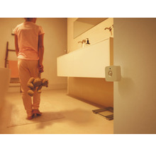 Détecteur de mouvement blanc carré fixé au mur dans une salle de bains tamisée avec une personne tenant un ours en peluche.