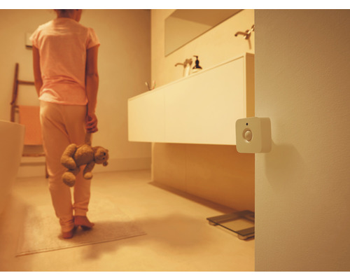 Détecteur de mouvement blanc carré fixé au mur dans une salle de bains tamisée avec une personne tenant un ours en peluche.