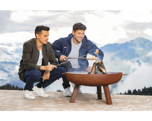 Zwei Personen grillen Stockbrot und Wurst über einer Feuerschale vor Bergkulisse.