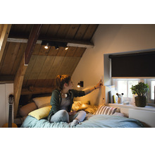 Chambre mansardée lumineuse avec plafonnier, stores et une femme actionnant un interrupteur.