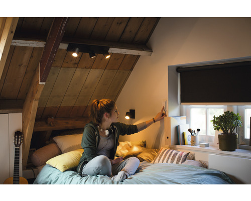 Chambre mansardée lumineuse avec plafonnier, stores et une femme actionnant un interrupteur.