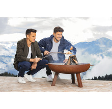 Deux hommes font griller de la pâte à pain et des saucisses au-dessus d''un brasero à l''extérieur, devant un paysage de montagne.