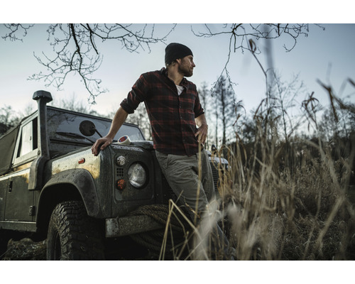 Mann in kariertem Hemd lehnt an einem Geländewagen in einer naturnahen Umgebung