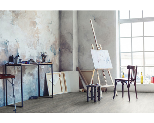 Atelier avec chevalet, toiles, table avec matériel de peinture et chaise devant une fenêtre.