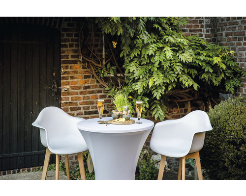 Scène extérieure avec une table haute, deux chaises, des boissons et des collations devant un mur de briques et des plantes.