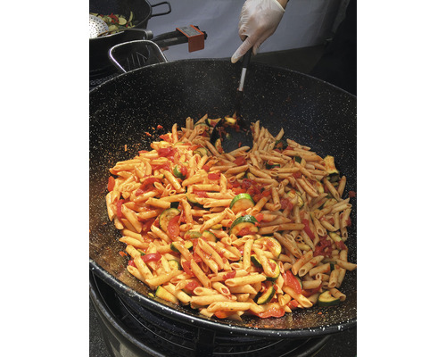 Une grande poêle contenant des penne, des courgettes et des tomates est remuée par une main avec une spatule.