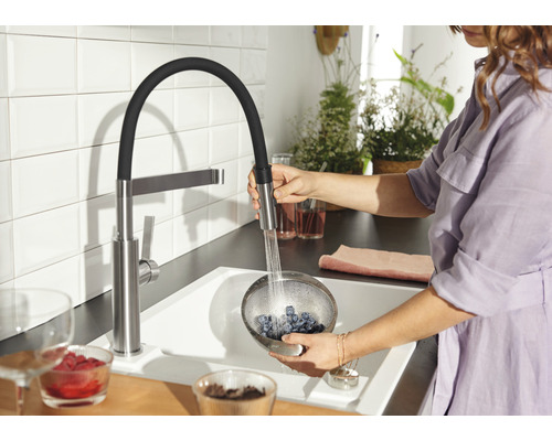 Une femme lave des myrtilles dans une passoire sous un robinet de cuisine avec douchette extractible dans une cuisine lumineuse.