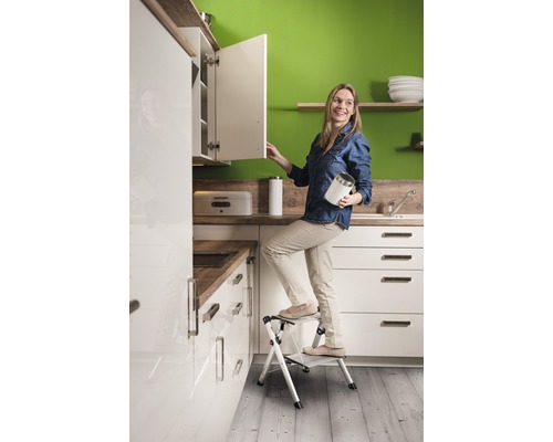 Femme sur un escabeau dans une cuisine avec des armoires blanches et des plans de travail imitation bois