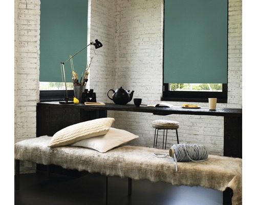 Helles Zimmer mit zwei Fenstern, die mit Rollos versehen sind, einer Bank und einem Schreibtisch mit dekorativen Objekten