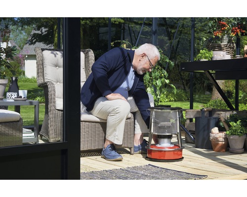 Un homme actionne un chauffage à pétrole dans une serre lumineuse avec des meubles et des plantes.