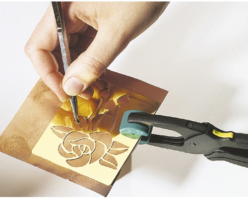 Une personne grave une rose dans une plaque de métal avec un outil de gravure, la plaque étant maintenue par une pince.