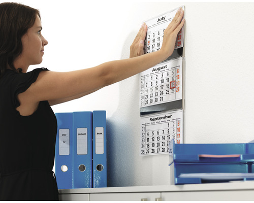 Une femme fixe un calendrier au mur dans un bureau à côté de classeurs et de corbeilles de rangement.