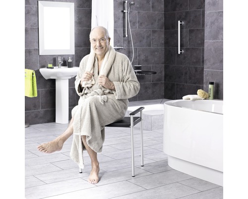 Un homme en peignoir est assis sur un siège de douche dans une salle de bains avec douche, lavabo et baignoire.