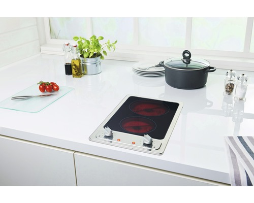 Küchenszene mit Kochfeld, Topf, Geschirr und Lebensmitteln auf einer weißen Arbeitsplatte.