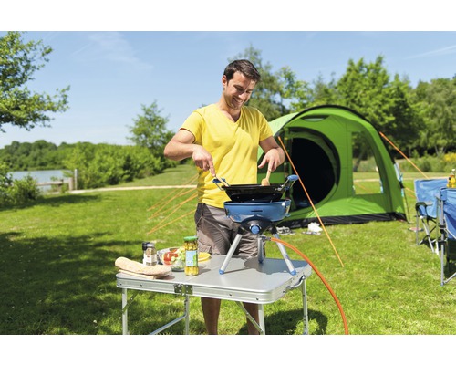 Mann kocht im Freien mit Campingkocher, Tisch und Zelt im Hintergrund