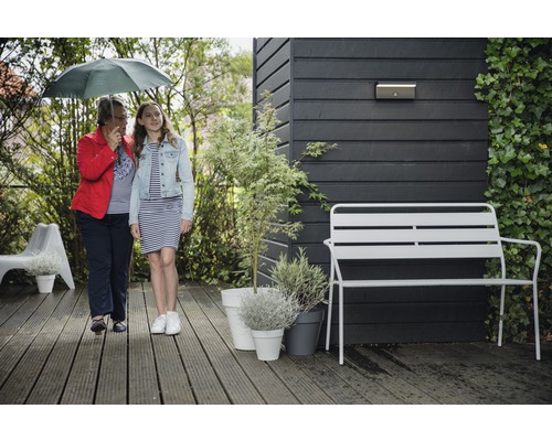 Gartenszene mit zwei Personen unter einem Regenschirm, einer Gartenbank, Pflanzen in Töpfen und einer Wandleuchte an einer Holzwand.