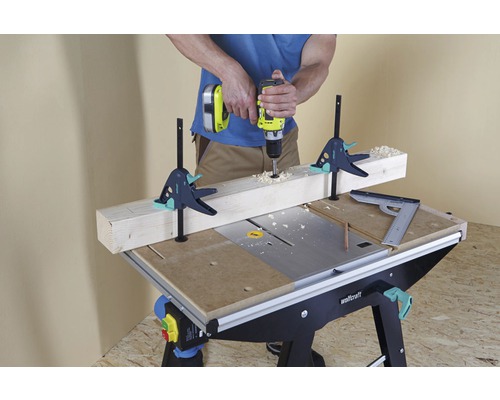 Handwerker bohrt mit einer Bohrmaschine ein Holzstück auf einer Werkbank, fixiert mit Schraubzwingen.