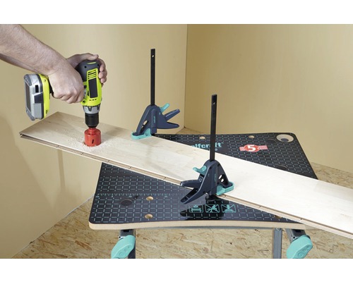 Eine Person bohrt mit einer Bohrmaschine ein Loch in ein Holzbrett auf einer Werkbank, die mit Schraubzwingen befestigt ist.