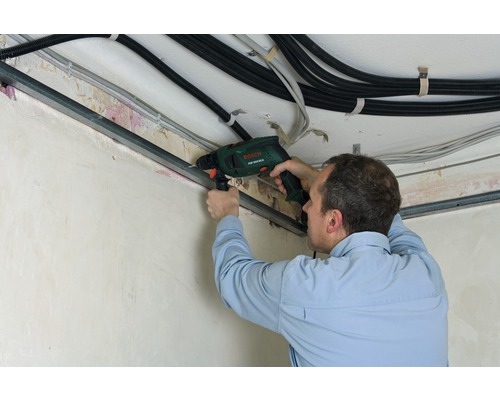 Un homme perce un rail métallique au plafond avec une perceuse à percussion. Des câbles sont également visibles.