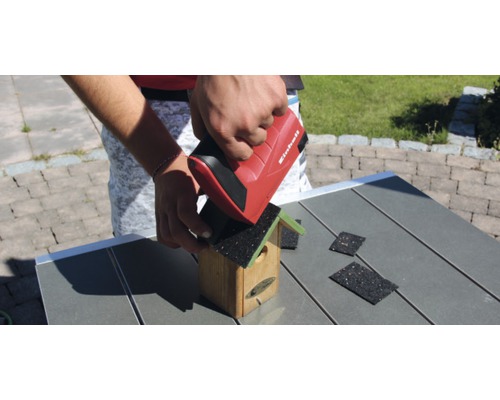 Eine Person befestigt mit einem roten Tacker von Einhell Dachpappe auf einem hölzernen Vogelhaus.