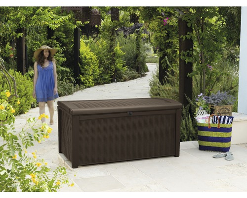 Braune Gartenbox aus Kunststoff auf einer Terrasse mit Frau im Hintergrund