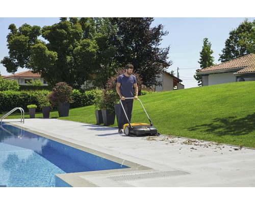 Un homme utilise une balayeuse pour nettoyer une terrasse à côté d''une piscine dans un jardin.