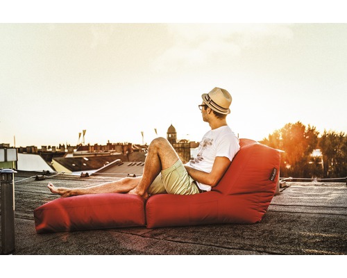 Mann mit Hut entspannt auf rotem Sitzsack mit Fussteil auf einem Dach