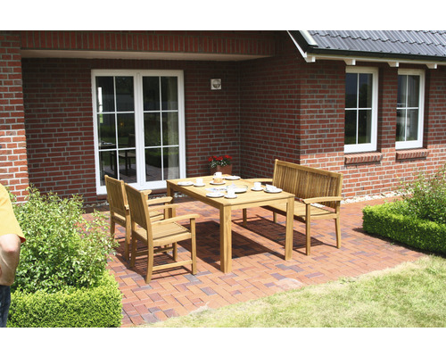 Scène de jardin avec une table à manger en bois et des chaises assorties sur une terrasse devant une maison en briques.