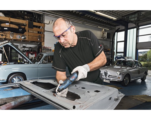 Un homme travaille avec un outil multifonction sur une portière de voiture dans un atelier avec des voitures anciennes.