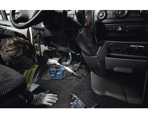 Un artisan nettoie l''intérieur d''un véhicule avec un aspirateur à main sans fil. Une visseuse à batterie est posée sur la moquette.