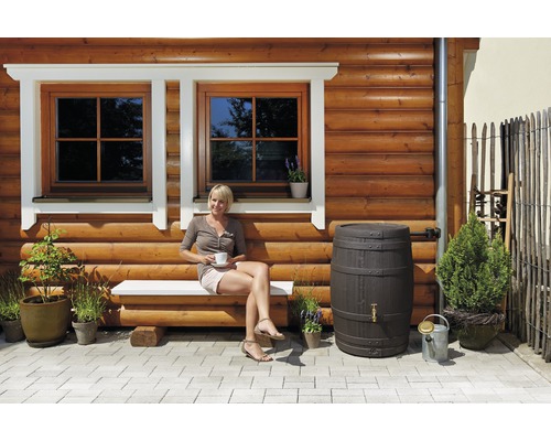Extérieur avec mur en bois, fenêtres, banc, femme assise, récupérateur d''eau de pluie et plantes