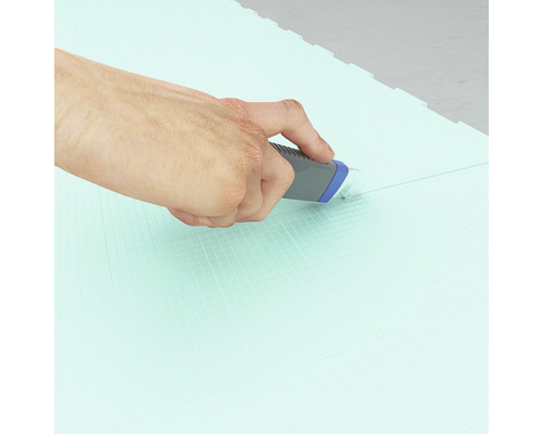 Une main coupe un tapis de sol vert avec un couteau universel.