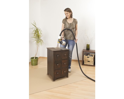 Femme vernissant un meuble en bois avec un système de pulvérisation de peinture, une feuille de protection de sol est posée