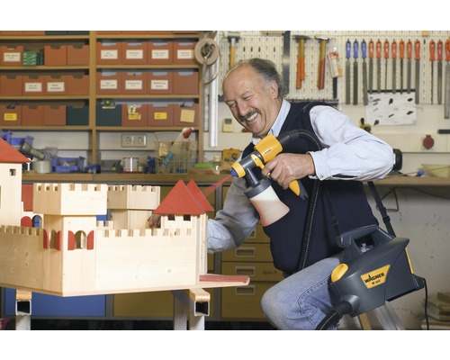 Un homme peint un château en bois avec un système de pulvérisation de peinture Wagner dans un atelier.