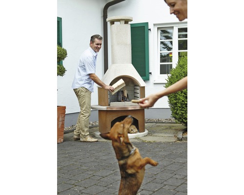 Scène avec un homme mettant du bois dans un barbecue de jardin pendant qu''une femme donne une friandise à un chien.