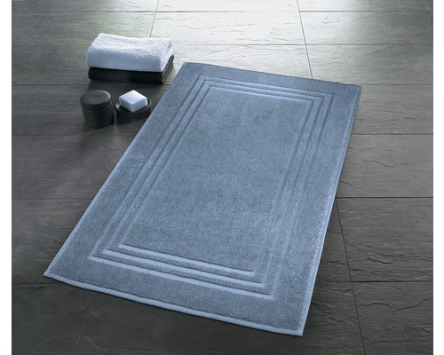 Rechteckige Badematte mit umlaufendem Rahmenmuster, daneben liegen Handtücher und Badutensilien