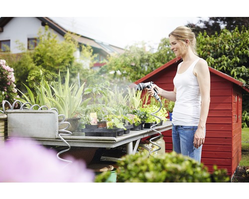 Une femme arrose des plantes avec un pistolet d''arrosage sur une table de jardin devant une cabane de jardin rouge.