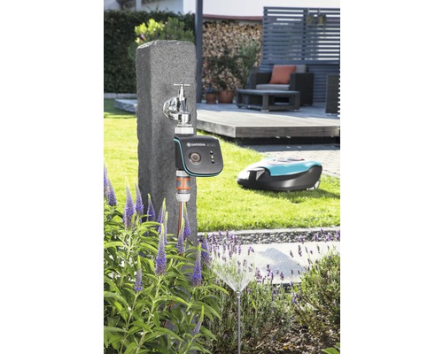 Scène avec un robinet, un programmateur d''arrosage Gardena, un arroseur de pelouse et un robot tondeuse dans le jardin.