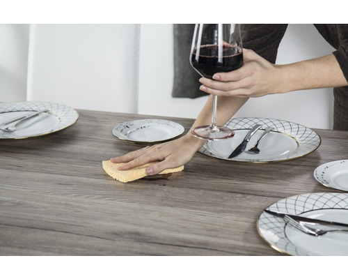 Scène avec table, couverts et une personne nettoyant une tache avec une éponge tout en tenant un verre de vin.