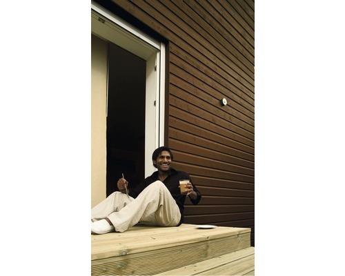 Mann sitzt auf einer Holzterrasse vor einem Haus mit Holzfassade.