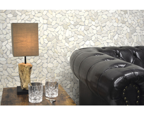 Scène avec une lampe de table sur une table en bois, deux verres, un ouvre-bouteille et un canapé en cuir devant un mur de galets.
