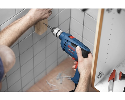 Une personne perce un trou dans une planche de bois dans une salle de bain avec une perceuse bleue.