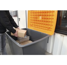 Une personne dépose des paquets dans une poubelle de recyclage grise avec un couvercle orange ouvert
